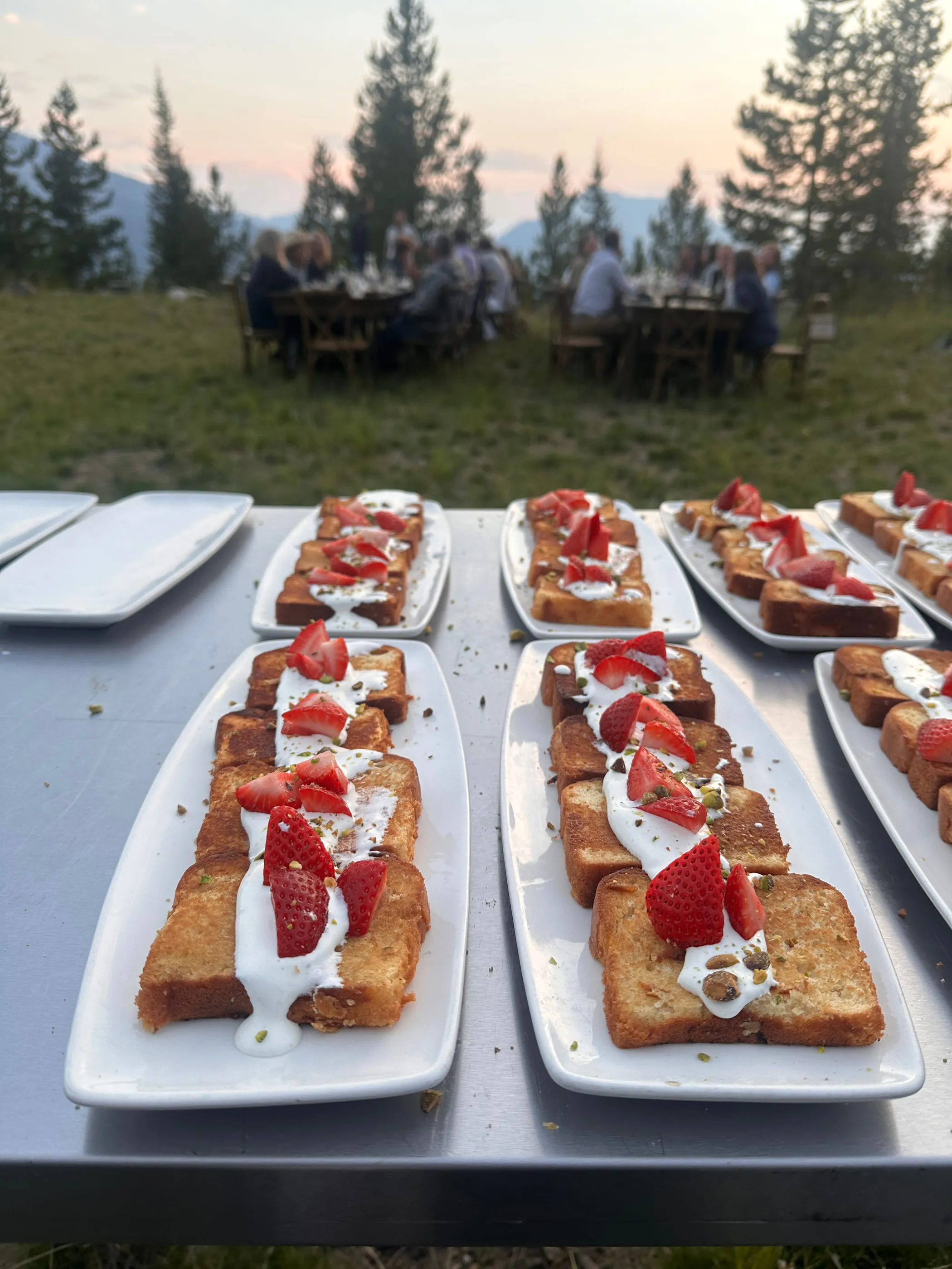 Brioche French toast with strawberries and cream plated along a long table, guests eating at communal tables in a mountain meadow at sunset behind.