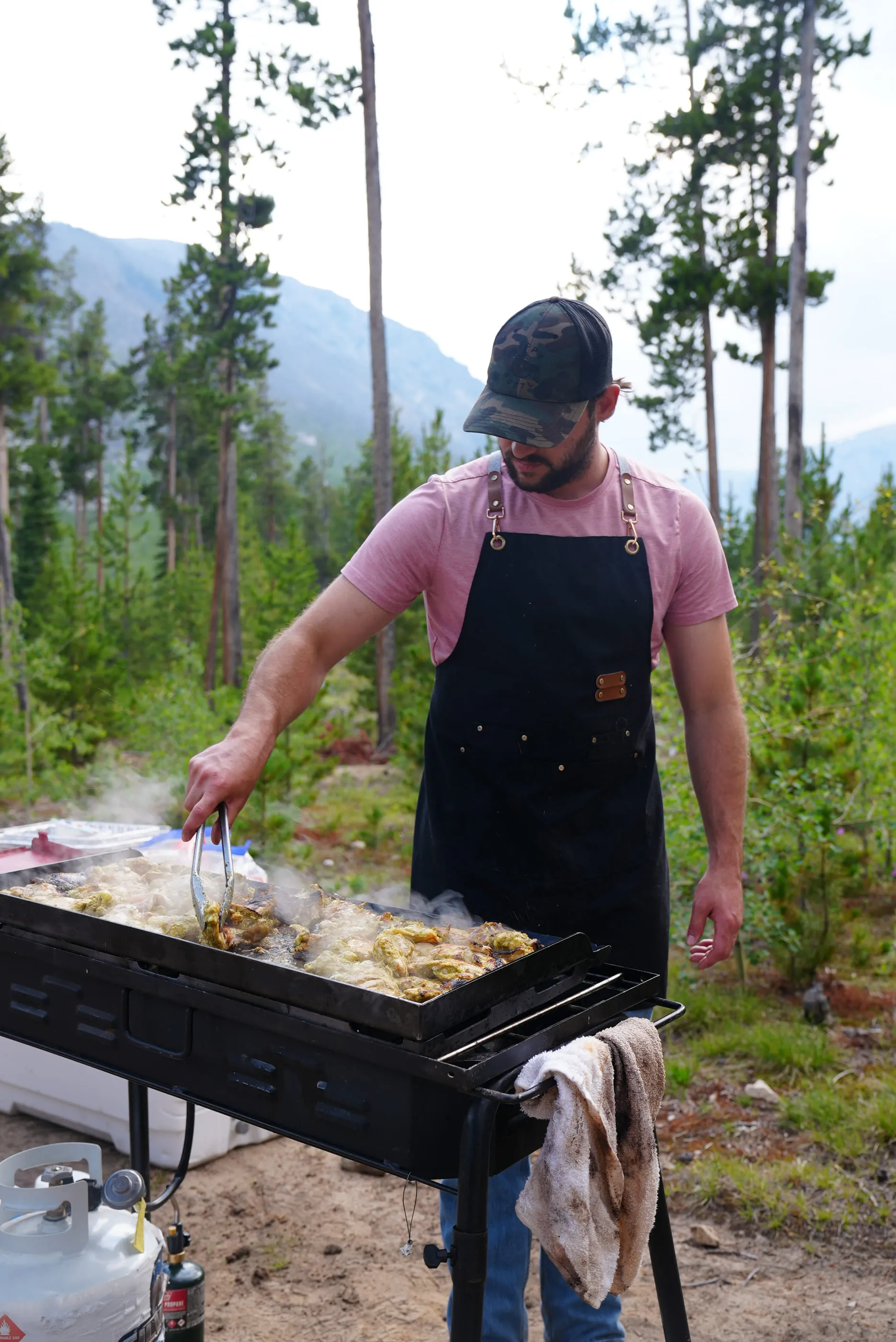 Nick working over a flat-top grill in camp, steam rising, pine forest and mountains behind.