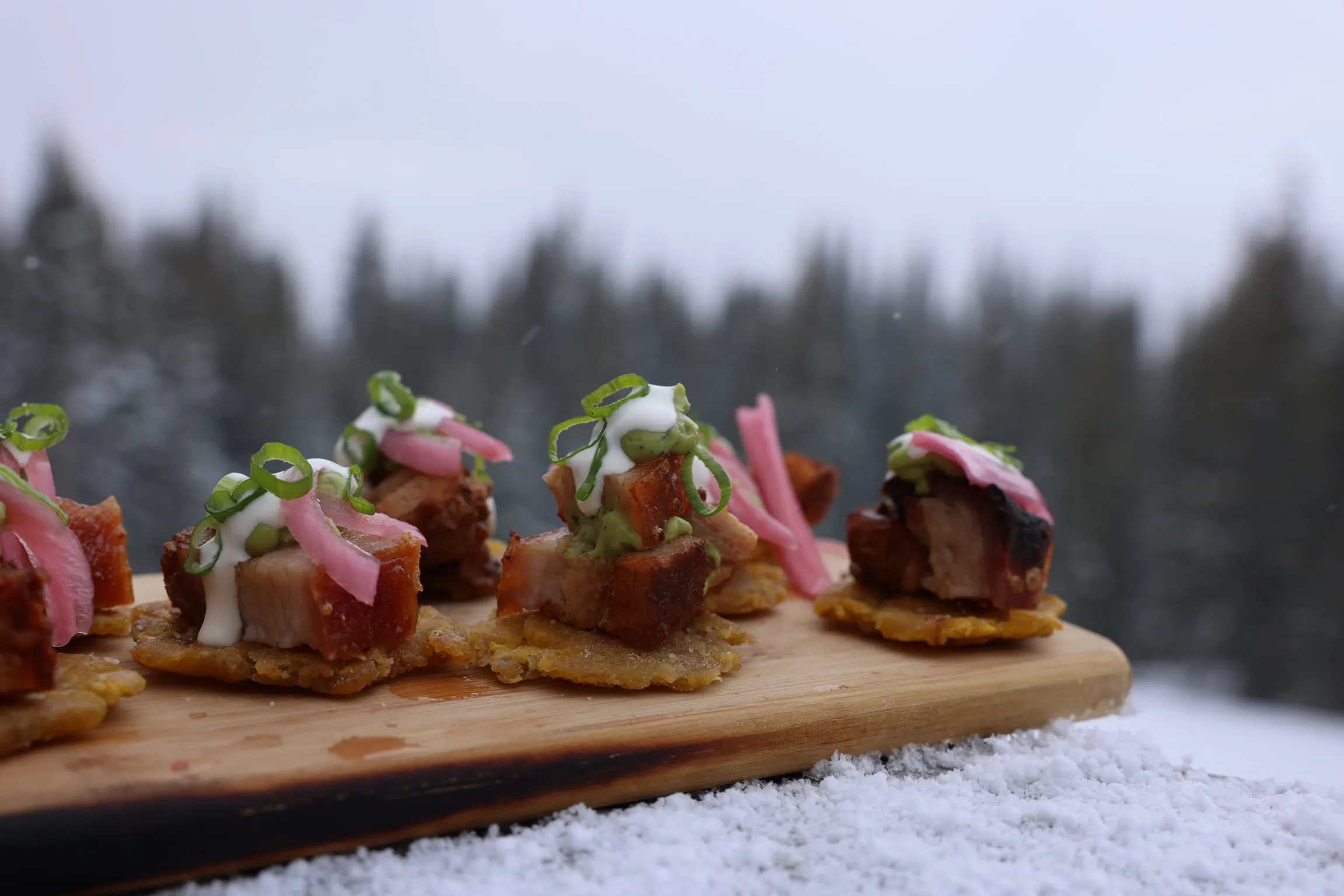 Pork belly bites with pickled onion and crema on a wooden board, a snow-covered treeline blurred behind.