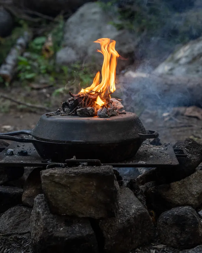 A cast-iron dutch oven on a rock fire ring, flames burning on the lid, deadfall and forest floor behind.