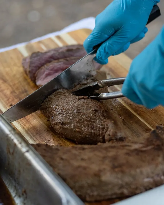 Gloved hands slicing a roasted cut of wild game on a wooden board at an outdoor prep station, a pan of jus alongside.